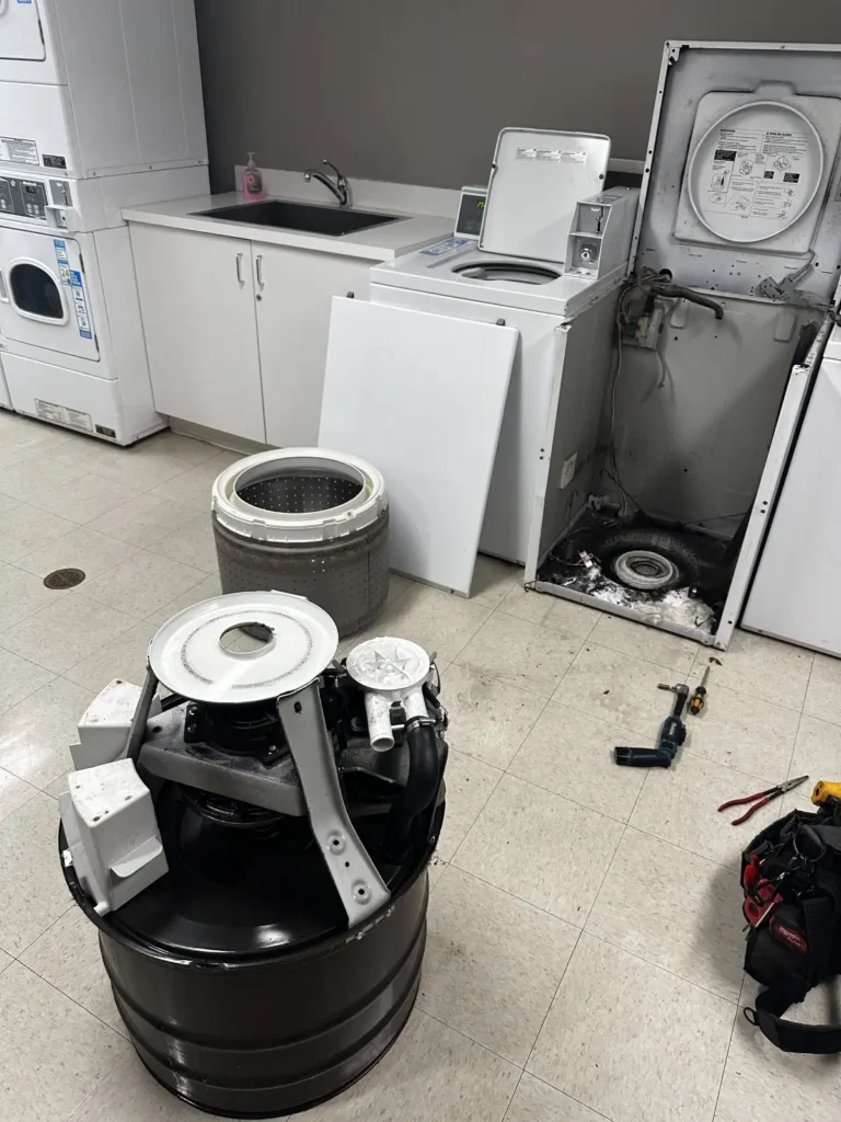 Disassembled top-load commercial washing machine with the inner tub, outer drum, and drive motor assembly separated on a laundry room floor