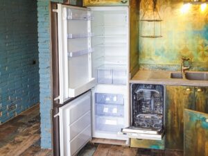 Empty built-in fridge freezer combo next to an open dishwasher under a kitchen counter.