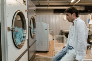 Man in a white shirt waiting by industrial washing machines running in a laundromat.